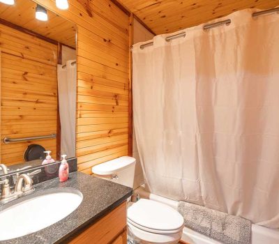 A small bathroom with wood-paneled walls and ceiling, a white sink with a black countertop, a toilet, a shower with a white curtain, a wall mirror, and bright lighting above the mirror.