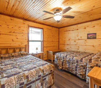 A cozy cabin bedroom with wood-paneled walls and ceiling, featuring two double beds with patchwork quilts, a window with blinds, a ceiling fan with a light, and wooden furniture including a nightstand and desk.