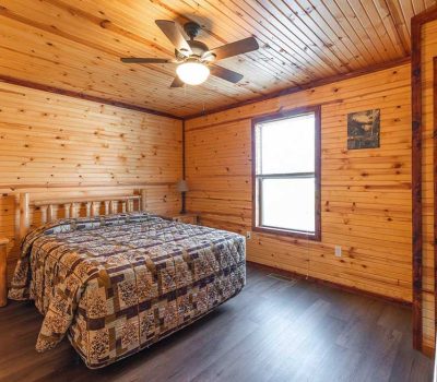 A cozy bedroom with wood-paneled walls and ceiling, a ceiling fan, a bed with a patchwork quilt, a bedside table, a window letting in natural light, a TV mounted in the corner, and a wooden door.