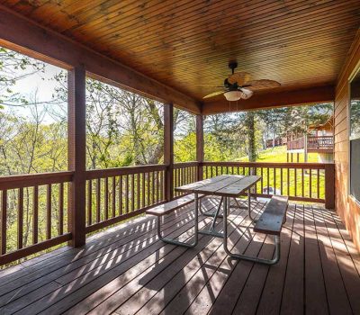 A covered wooden porch with a picnic table, benches, a metal chair, ceiling fan, and railing, overlooking green trees and grass on a sunny day.