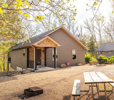 A small brown cabin with a covered porch sits among trees, with a picnic table and fire pit in the foreground. A "For Sale" sign is visible near the front door, and sunlight filters through the branches.