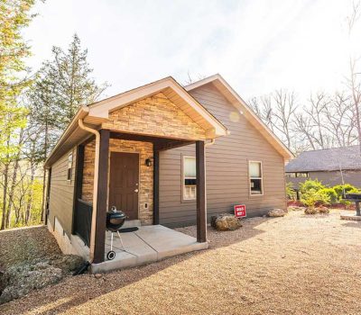 A small brown cabin with a covered front porch sits on a gravel lot surrounded by trees. The sun is shining brightly, casting shadows, and a grill is near the front door. A red sign is visible by the side of the house.