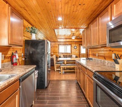 A cozy log cabin kitchen with wood-paneled walls and ceiling, stainless steel appliances, brown cabinets, a double sink, and a dining area with a wooden table and benches in the background.