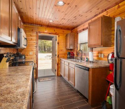 A cozy kitchen with wooden walls and ceiling, brown cabinets, granite countertops, stainless steel appliances, and an open door leading outside to a picnic table in a wooded area.