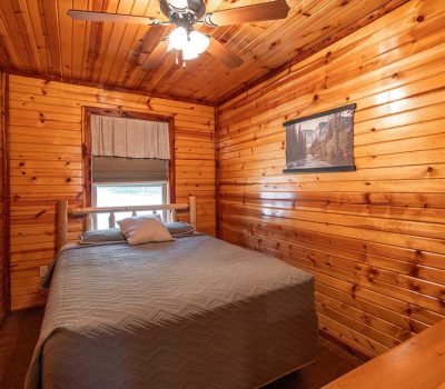 A cozy bedroom with wood-paneled walls and ceiling, featuring a ceiling fan, a bed with a gray quilt and pillows, a window with a shade, and a landscape picture hanging on the wall.