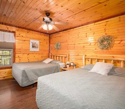 A cozy cabin bedroom with wood-paneled walls and ceiling, two beds with gray bedding, a ceiling fan with lights, a wall-mounted TV, and a window overlooking a porch. Wreaths and animal art decorate the walls.