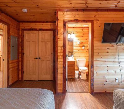 A cozy wood-paneled cabin bedroom with a wall-mounted TV, visible bathroom with toilet and sink, closed double doors, and parts of two beds with quilted blankets.
