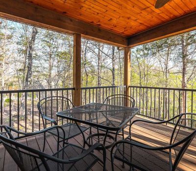 A covered wooden deck with a square metal table and four matching chairs overlooks a wooded area with tall trees and sunlight filtering through the branches.