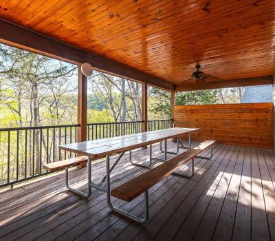 A covered wooden deck with a long picnic table and benches, surrounded by a black railing, overlooks green trees and has a ceiling fan on the wood-paneled ceiling.