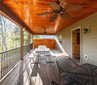 A covered wooden porch with a ceiling fan, picnic tables, metal chairs, and a railing, surrounded by trees. The porch has a wooden ceiling and siding, with sunlight streaming in.