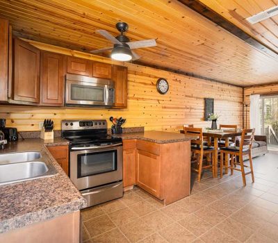 A cozy kitchen with wood-paneled walls and ceiling, stainless steel appliances, brown cabinets, a double sink, and a dining area with a table and chairs near large windows letting in natural light.