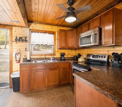 Rustic kitchen with wooden cabinets and walls, stainless steel appliances, a window with a curtain, brown countertops, and a ceiling fan. The door is open, showing a view of trees and rocks outside.