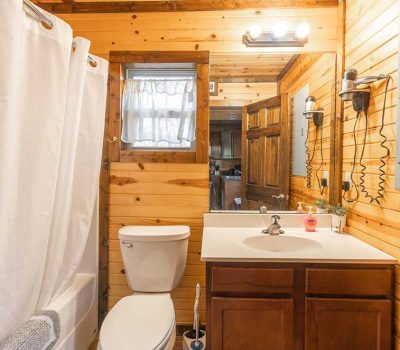 A rustic bathroom with wood-paneled walls, a white toilet, a bathtub with a white shower curtain, a sink with a vanity, a mirror, and wall-mounted lights above the mirror. A window lets in natural light.