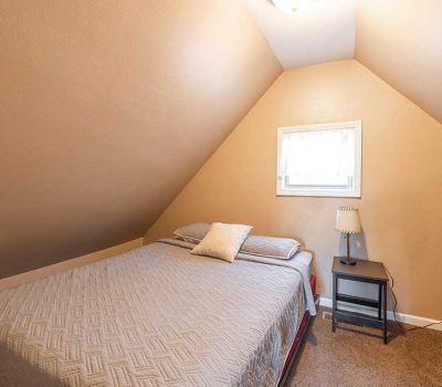 A small bedroom with sloped ceilings, featuring a neatly made bed with a gray quilt and pillows, a small window above the bed, and a black nightstand with a lamp, all set on a brown carpeted floor.
