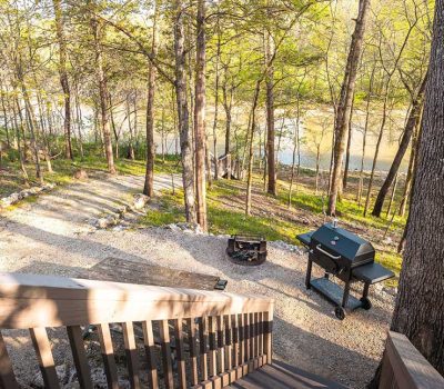 Wooden steps lead down to a gravel path beside a fire pit and outdoor grill, surrounded by tall trees and overlooking a river in a lush, green forest setting on a sunny day.
