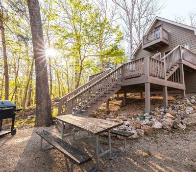 A wooden cabin on a rocky slope with a large staircase, surrounded by trees. In front, there is a picnic table and a barbecue grill, with sunlight streaming through the forest.