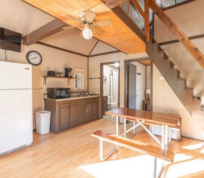 A bright, rustic kitchen with a white refrigerator, wooden cabinets, microwave, wall clock, and a rectangular wooden table with benches. Wooden stairs lead to a loft area; sunlight streams in from the right.