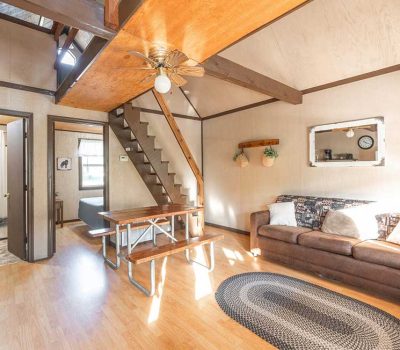Bright, cozy living room with a brown sofa, pillows, wall art, a wooden table, and a small staircase leading to a loft. Wooden beams, a ceiling fan, and natural light enhance the warm, inviting atmosphere.