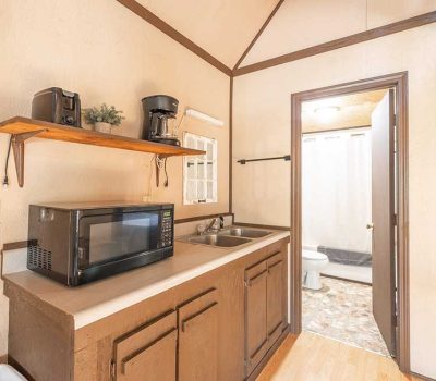 A small kitchenette with a microwave, sink, and brown cabinets. A wooden shelf above holds a coffee maker, a plant, and a clock on the wall. A bathroom with a toilet and shower is visible through an open door.