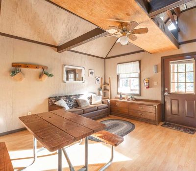 A cozy, sunlit cabin interior with wooden floors, a brown couch, a picnic-style dining table, a wooden dresser, and windows with white curtains. A ceiling fan and loft ladder are also visible.