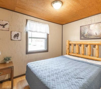 A cozy bedroom with a rustic wooden bed, light blue quilt, wooden nightstand with a lamp and plant, wall art featuring animals, and a window with a white valance letting in natural light.