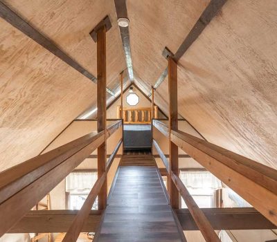A narrow wooden loft walkway with railings leads to a cozy bed under a pitched ceiling in a rustic attic room, with natural light streaming through windows at the end and below.