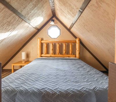 A cozy attic bedroom with wooden beams, a log-style bed frame, a gray quilted bedspread, a small bedside table with a lamp, and a small octagonal window on the far wall letting in natural light.