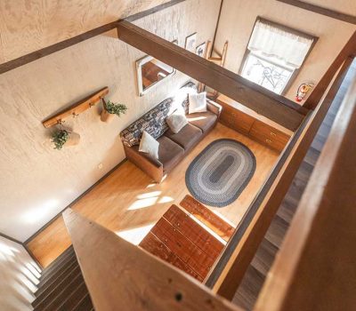 A cozy living room seen from above, featuring a brown sofa with pillows, a wooden chest, a woven oval rug, and natural light streaming through a window with white curtains. Wooden stairs and decor are also visible.