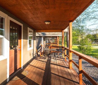 A wooden porch with a table and chairs overlooks a green yard with trees. Sunlight casts shadows on the porch and highlights the natural outdoor scenery.