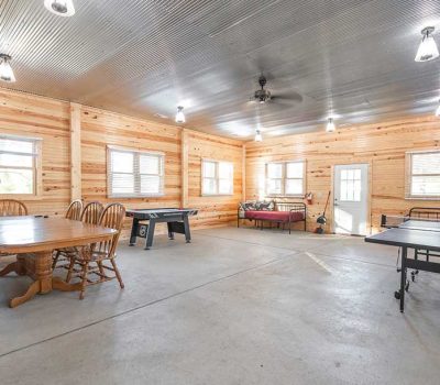 A spacious recreation room with wood-paneled walls, a large table with chairs, a table tennis setup, a foosball table, and daybeds, all under a corrugated metal ceiling with several lights.
