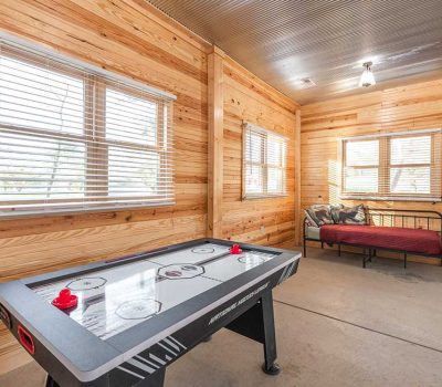 A cozy room with wood-paneled walls features an air hockey table, a daybed with pillows, large windows with blinds, and cleaning supplies in the corner. Natural light fills the space.