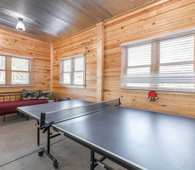 A bright room with wood-paneled walls, a black ping pong table in the center, a daybed with burgundy bedding and pillows, and three windows with blinds letting in natural light.