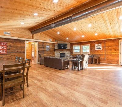 Spacious wooden lodge interior with vaulted ceiling, large dining table and chairs, cozy seating area with sofas, TV, and fireplace. Sunlight streams through glass doors. A red “Dining Lodge” sign hangs on the wall.