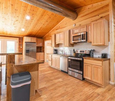 Modern kitchen with wooden floors, walls, and ceiling, stainless steel appliances, light wood cabinets, a small island with stools, and a trash can in the foreground. Double doors provide natural light in the background.