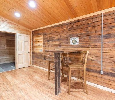 A rustic room with wooden walls, floor, and ceiling, featuring a high wooden table and two chairs. There is a door leading to another room and a wall-mounted sign with visible wiring along the wall.