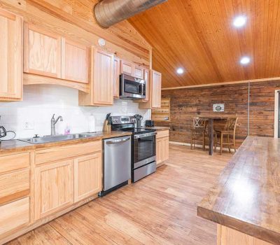 A spacious kitchen with light wood cabinets, stainless steel appliances, wood floors, and a wooden ceiling. There is a dining area in the background and bright ceiling lights illuminate the space.