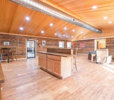 Spacious rustic kitchen and living area with wood floors, wooden ceiling, exposed ductwork, kitchen island, cabinets, dining table, and a stone fireplace in a cozy, well-lit open layout.
