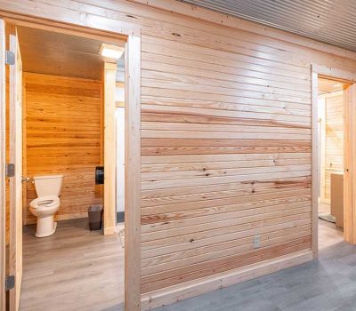 A hallway with light wooden walls and gray flooring, featuring open doors leading to restrooms; one bathroom has a visible toilet and trash bin. A fire extinguisher is mounted on the wall.
