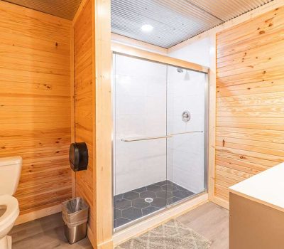 A modern bathroom with wood-paneled walls, a white toilet, a small trash can, a walk-in shower with glass doors, and a white sink vanity on the right.