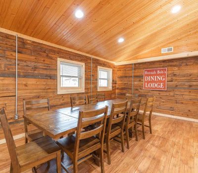 A rustic dining room with wooden walls, ceiling, and floor, featuring a long wooden table with eight chairs. A sign on the wall reads “Huzzah Bob’s Dining Lodge.” Two windows let in natural light.