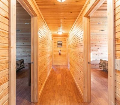 A bright hallway with wooden walls, floors, and ceiling, leading to two open doorways on either side, each revealing a bedroom with a bed visible. A picture hangs on the wall at the end of the hallway.