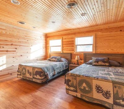 A cozy cabin bedroom with wood-paneled walls and ceiling, two beds with nature-themed quilts, a small nightstand with a lamp between them, and sunlight streaming through two windows.