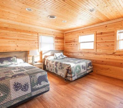 A cozy bedroom with wood-paneled walls and ceiling, featuring two beds with nature-themed quilts, a bedside table with a lamp, and three windows letting in natural light.