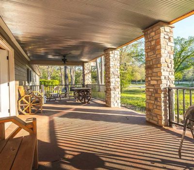 A spacious covered porch with stone columns, wooden bench, grill, and outdoor tables and chairs, overlooking a grassy yard with trees on a sunny day. Shadows from the railing create patterns on the floor.