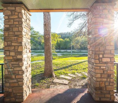 View from a covered porch with stone columns, looking out onto a sunny lawn and trees, with bright sunlight streaming through the scene. A pathway of stepping stones leads away from the porch.