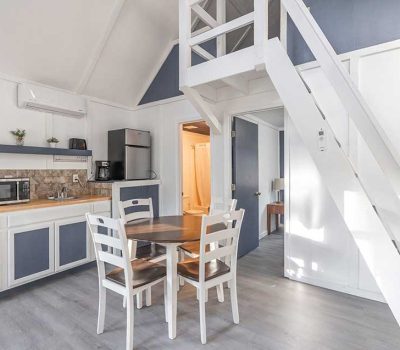 A bright, modern kitchen and dining area with white furniture, gray floors, a round table with four chairs, a microwave, a small fridge, and a staircase leading to a loft. Sunlight streams through a window.