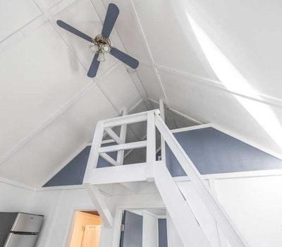 A bright white loft with a slanted ceiling, blue and white walls, a small staircase leading up, and a ceiling fan. Part of a refrigerator and a split air conditioner are visible below.