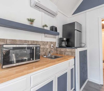 Small kitchen with a wooden countertop, microwave, sink, coffee maker, compact fridge, and open shelf with plants. White cabinets and walls, gray accents, and a view into a bathroom with a shower curtain.