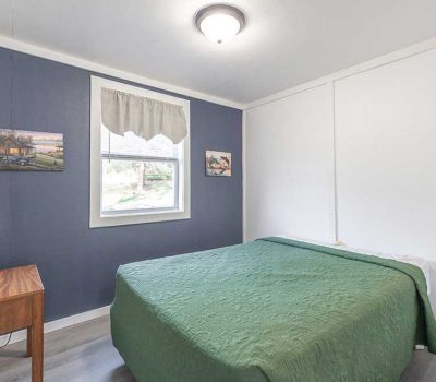 A small bedroom with a double bed covered in a green quilt, a wooden nightstand with a lamp, blue accent wall with two paintings, and a window letting in natural light.