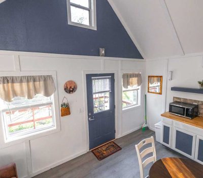 Interior of a small, cozy kitchen with white and blue walls, a door with a window, two curtained windows, a counter with a microwave, a sink, and a round dining table with chairs. A broom and wall decor are visible.